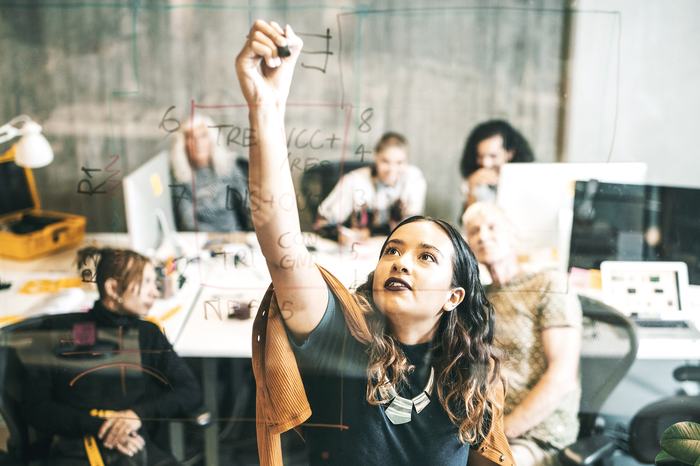 Young woman drawing on whiteboard in team meeting while others look on and discuss.